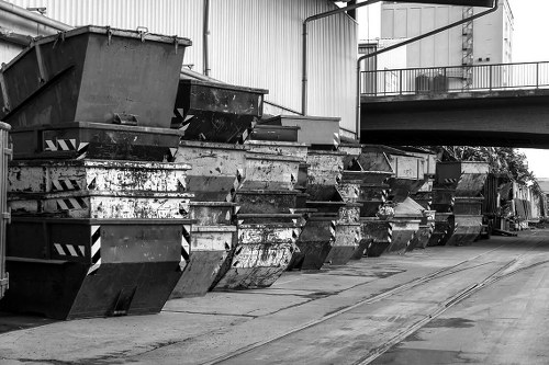 A row of large metal skips positioned along a paved outdoor area, likely part of a commercial waste collection site in Southwark. The skips are constructed from heavy-duty steel with a corrugated texture and are in varying shades of dark grey and black, some showing signs of rust and weathering. They are aligned side by side, with the open tops facing upward, some filled with debris or waste material. Behind the skips, there is a corrugated metal building with vertical panels and a metal railing on an elevated platform. The environment appears to be an industrial or warehouse district, with concrete pavement extending beneath the skips. The setting is well-lit, with natural daylight illuminating the scene and shadows cast by the skips and surrounding structures. The image reflects a typical outdoor waste management area that a company like Commercial Waste Removal Southwark might service, supporting efficient rubbish removal in the local area, which includes the Southwark postcode district.