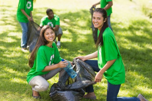 A group of young volunteers wearing matching bright green t-shirts with white recycling symbols participate in a community litter pick in a grassy park area. In the foreground, two women are kneeling on the ground, smiling at the camera while holding a large black plastic rubbish bag filled with collected waste. One of them is holding a plastic bottle and both are wearing gloves for safety. In the background, other volunteers can be seen standing and actively collecting trash, with some holding additional black plastic bags. The scene is set outdoors in a well-lit, sunny environment with lush green trees and grass, indicative of an organized environmental cleanup event focused on waste collection and community involvement. Commercial Waste Removal Southwark offers rubbish removal services in the Southwark area, supporting local environmental efforts across London.