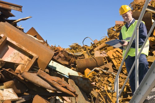 A male waste management technician wearing a yellow safety helmet and high-visibility vest is standing on a metal staircase at an industrial site in Southwark, inspecting a large pile of rusted metal debris. The debris consists of various steel panels, beams, and scrap metal with a rough, weathered texture and reddish-brown rusted surfaces. In the background, clear blue sky is visible above the noisy, outdoor setting. The technician is holding a clipboard and appears to be assessing the waste materials as part of a commercial rubbish removal operation, typical for services provided by Commercial Waste Removal Southwark, supporting efficient waste clearance in the Southwark area.