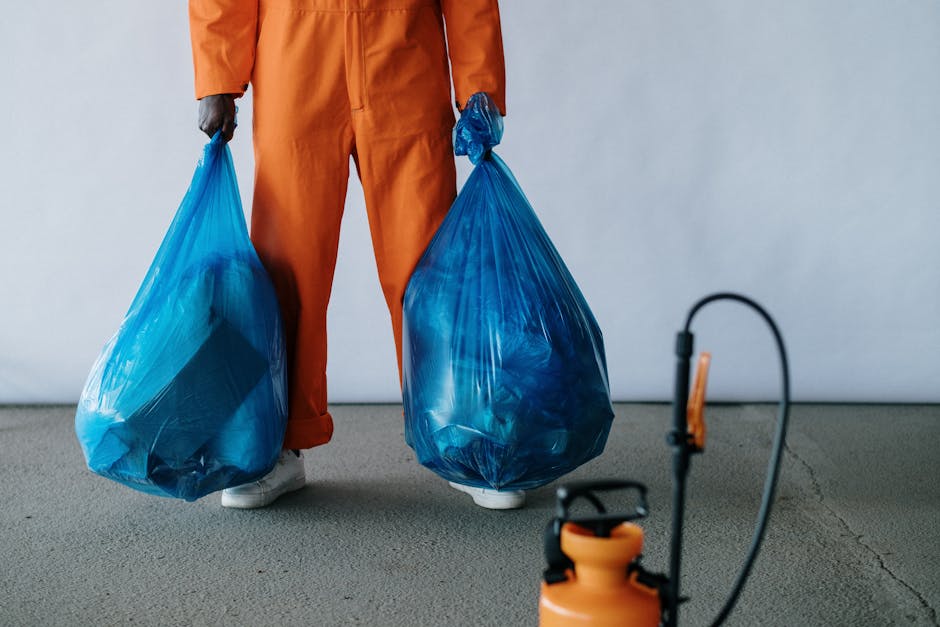 A waste collection worker wearing a high-visibility yellow vest and blue gloves operates a large, white waste collection truck on a city street during dusk. The truck's rear compartment is partially o
