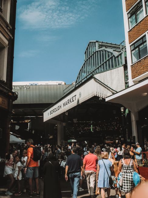 A covered outdoor area at Borough Market in SE1, featuring black metal support columns and a curved corrugated roof with a light beige finish. In the background, a red metal railings section encloses
