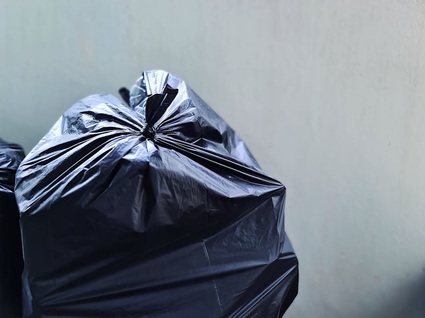 Three large black plastic garbage bags filled with waste materials are positioned on the edge of a paved street in front of a black metal railing and a wooden fence, with dense foliage visible behind