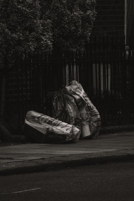 Two large black garbage bags filled with waste materials are positioned on a sidewalk in front of a black metal fence, with dense foliage visible behind the fence. The bags appear to be made of heavy-