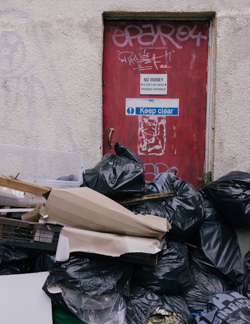 Two large black garbage bags filled with waste materials are positioned on a sidewalk in front of a black metal fence, with dense foliage visible behind the fence. The bags appear to be made of heavy-