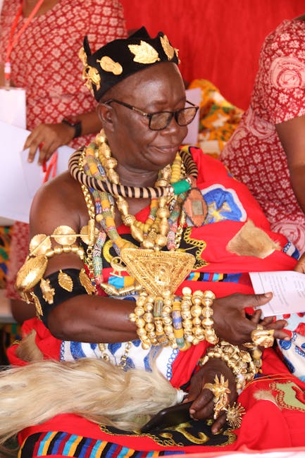 A woman wearing traditional African attire, dressed in vibrant red, with intricate gold and multi-coloured beadwork adorning her neck, arms, and headpiece. She is seated, holding a piece of paper in h